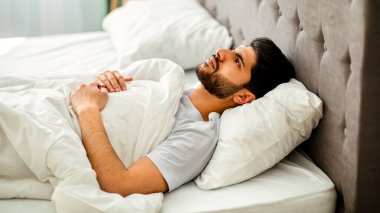 Uykusuzluk. Depresyondaki Arap adam uyuyamıyor, yatakta uzanıyor, yukarı bakıyor ve uzayı kopyalıyor. Uykusuzluk, akıl sağlığı sorunu, erkek depresyonu kavramı.