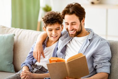- Boş zaman. Mutlu baba ve oğul kitap okuyor ve gülümsüyor, oturma odasındaki rahat kanepede dinleniyorlar. Ebeveynlik ve aile ilişkisi, hafta sonunu evde birlikte geçirmek.