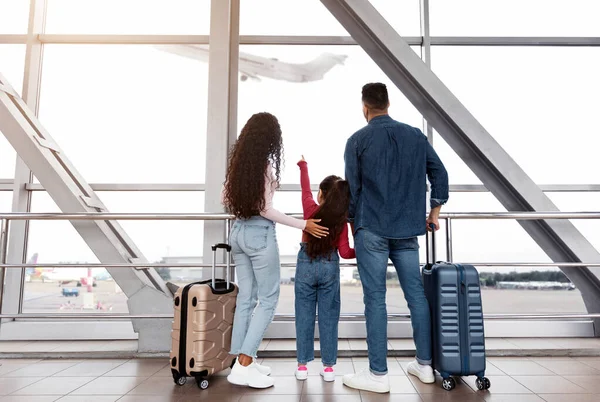 Havalimanı terminalinde ailesiyle birlikte duran, pencereden dışarı bakan, arka manzaralı sevimli küçük kız. Üç kişilik Orta Doğulu bir aile uçağa binmek için bekliyor, birlikte tatile çıkmaya hazır.