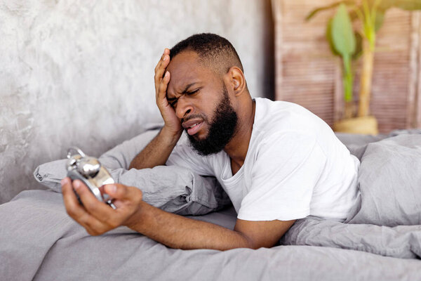Потрясенный чернокожий мужчина смотрит на будильник, лежит дома в постели, трогает голову, опаздывает на работу. Нервный афроамериканец, проспавший, с трудом просыпающийся утром