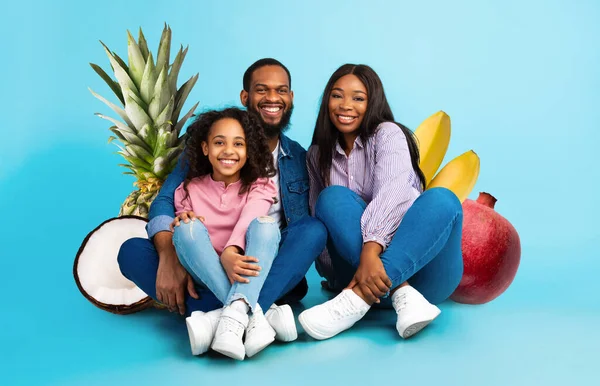 Neşeli bir aile, ananas, hindistan cevizi ve nar gibi renkli meyvelerle çevrili mavi bir arka plana karşı birlikte oturur. Sağlığı ve besinleri gülümseyerek kutlarlar..