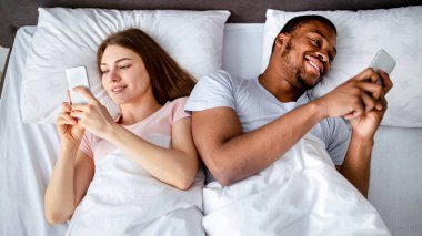 Gadget bağımlılığı ve ilişki sorunları. Akıllı telefonları yatakta uzanan, birbirlerini görmezden gelen, online oyunlar oynayan, sosyal medyayı karıştıran, üst görünümü olan bin yıllık ırklar arası bir çift.