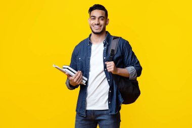Sırt çantalı ve bir sürü kitabı olan zeki Arap öğrenci kameraya gülümsüyor, sarı stüdyo arka planında reklam için alanı kopyalıyor. Eğitim, üniversite, üniversite, öğrenim, ders kavramı