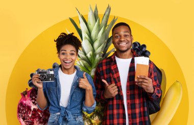 İki neşeli kişi çeşitli meyvelerle dolu renkli bir arka plan önünde poz veriyor. Ellerinde bir kamera ve seyahat belgeleri var. Sağlıklı bir yaşam tarzını ve keşiflerin zevkini kucaklıyorlar..