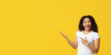 Şuna bir bak. Aşırı neşeli Afro-Amerikalı kadın boş yeri işaret ediyor, reklam için fotokopi alanını gösteriyor, sarı arka planda tek başına duruyor, boş alanı olan bir panorama.