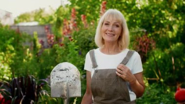 Son sınıf öğrencisi bir kadın neşe içinde, bol bir bahçede duruyor, tulum giyiyor ve elinde bir kürek, güneşli gökyüzünün altında renkli bitkiler ve çiçeklerle çevrili, neşeli bir bahçe deneyimini yansıtıyor..