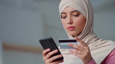 Başörtüsü takan genç Müslüman bir kadın elinde bir kredi kartı tutarken akıllı telefonuna odaklanıyor ve modern evinde online alışveriş yapıyor..