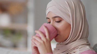 Tesettürlü Müslüman kadın evinde rahat bir şekilde oturmuş pembe bardaktan çay yudumluyor, rahatlama ve memnuniyet anına dalmış, modern bir yaşam tarzı sergiliyor..