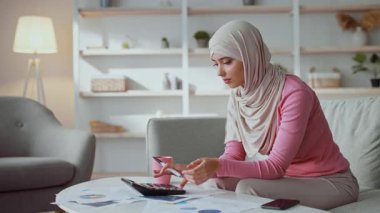 Başörtülü genç bir kadın kanepeye oturur ve hesap makinesine konsantre olur. Kağıtlar ve çizelgeler masaya serildi. Aile bütçesini saymakla meşgul olduğunu gösteriyor..