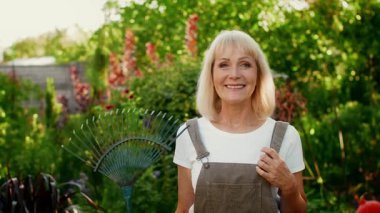Neşeli yaşlı bir kadın çiçeklerle ve yeşilliklerle dolu canlı bir bahçede elinde tırmıkla duruyor ve sıcak güneş ışığı altında bahçe işlerine olan aşkını gösteriyor..