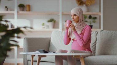 Orta Doğulu, tesettürlü hamile bir kadın koltuğunda oturup sıcak bir içki yudumluyor. Rahat bir şekilde giyinmiş ve rahat bir ev ortamı ile çevrili, sakin bir yaşam tarzını yansıtıyor..