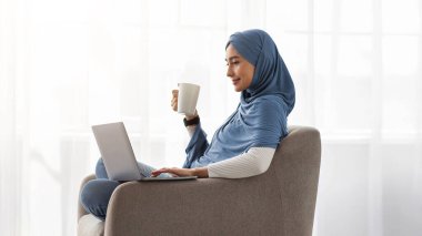 Rahat bir iş yeri, huzur içinde yat, tembellik yap, video izle, film izle. Minimalist oturma odasında dizüstü bilgisayarlı tesettürlü mutlu bin yıllık Arap bayan pencere arkasında çay içiyor, profil çıkarıyor.