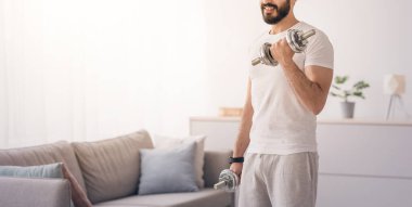 Covid-19 karantina ve boş zaman aktiviteleri, spor ve evde sağlıklı bir yaşam tarzı için egzersiz. Gülümseyen yakışıklı Arap sakallı milenyum sakallı adam oturma odasının arka planında dambıllar kaldırıyor.
