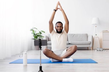 Gerçekten sakin, sakallı, beyaz spor giyimli, Lotus pozisyonundaki Afro-Amerikan bir adam oturma odasının içindeki web kamerasından video kaydediyor. Spor salonu evde. Çevrimiçi fitness kursu, COVID-19 için kendi kendine bakım
