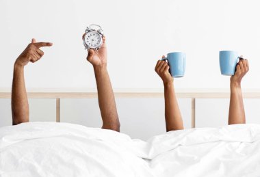 Hands of millennial african american man and lady stick out from under blanket on bed in white bedroom interior with coffee cups and alarm clock. Good morning and drink for energy, time to wake up