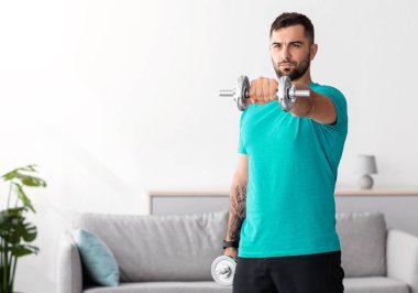 Mavi tişörtlü ciddi bir Avrupalı atlet minimalist oturma odasının iç kısmında dambıl kaldırıyor. Covid-19 salgını, spor yaşam tarzı, güç egzersizleri ve vücut bakımı sırasında yeni normaller