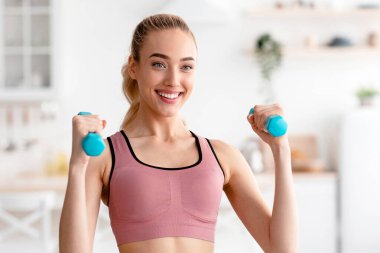 Mutlu sporcu evde çalışır, eller için egzersiz yapar ve COVID-19 kilitlenirken formda kalır. Gülümseyen genç, çekici, sarışın kadın spor kıyafetleri içinde halterleri kaldırıyor.