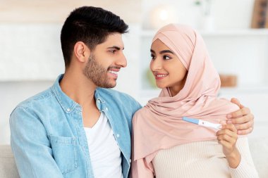Uzun zamandır beklenen hamilelik. Mutlu Arap ailesi pozitif hamilelik testi tutuyor koltukta oturuyor ve birbirlerine gülümsüyorlar. Genç Müslüman çift bebek bekliyor. Ebeveyn konsepti.
