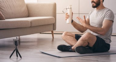 Sağlık hizmeti, spor blogu, online konferans, uzaktan giysi. Mutlu Arap sakallı genç 30 'lu yaşlarda bir adam paspasın üzerinde oturur, oturma odasının içinde el ele su şişesini gösterir, yerdeki telefon kamerasına bakar.