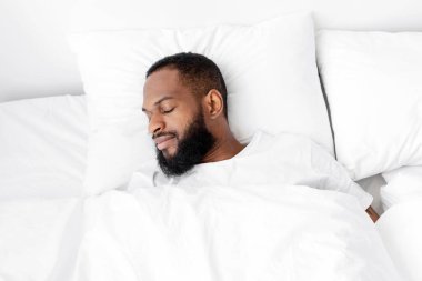 Sakin bin yıllık siyah sakallı erkek yatakta uyuyor, yatak odasında beyaz yorganı olan yumuşak yastıklı hafta sonları dinleniyor ve tatilin tadını çıkarıyor, boş alan. Sağlıklı uyku, sağlık hizmetleri ve evde dinlenme.
