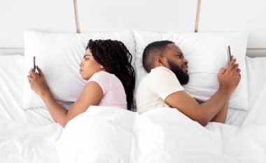Sad young black guy and lady lie back to back, looks at smartphones on bed in bedroom interior. Relationship problems, deception and treason, addiction of technology, online games and social networks