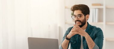 Ev ofisteki dizüstü bilgisayarda çalışan yakışıklı Arap girişimci, iş yerinde oturuyor, boş yer, panorama. Bilgisayar ekranına bakan adam, uzak işten zevk alıyor