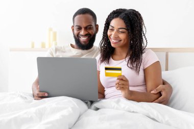 Smiling millennial african american guy with computer and lady with credit card shopping online in comfortable bed in bedroom. New normal and modern shopping in cozy home during covid-19 lockdown