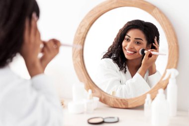 Günlük makyaj, güzellik dersleri, cilt bakımı ve kozmetik ürünleri. Beyaz önlüklü, gülümseyen Afro-Amerikan bayan makyaj malzemeleri ve yuvarlak ayna ile yatak odasındaki fırçayla yüzüne pudra sürüyor.