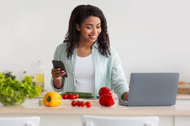 Modern cihazlar, uzaktan çalışma, yeni yemek tarifi ve yemek blogu için yemek pişirme. Gülümseyen Afro-Amerikan bayan dizüstü bilgisayarda yazı yazıyor, mutfak içlerinde akıllı telefon kullanıyor ve masasında parlak sebzeler var.
