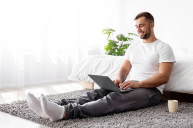 Gülümseyen beyaz, yakışıklı, kaslı bir adam yatak odasında dizüstü bilgisayarda oturuyor. İçi kahve, güneş ışığı, boş yer. Sağlık hizmetleri, serbest çalışma, ev ve iş blogu