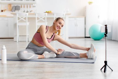 Koç uzaktan, fitness blogcusu evde çalışıyor, covid-19 tecrit. Spor kıyafetli mutlu genç bir sarışın bacak arası esneme hareketleri yapıyor, oturma odasındaki web kamerasına bakıyor, içinde bir şişe su var.