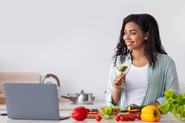 Genç, güzel bir kadın mutfakta elinde bir bardak şarapla yemek pişiriyor arkadaşlarıyla sohbet ediyor, internetten arıyor. Gülümseyen Afrikalı Amerikalı kadın yemek keser ve şarap içer, masada dizüstü bilgisayar ve sebzelerle