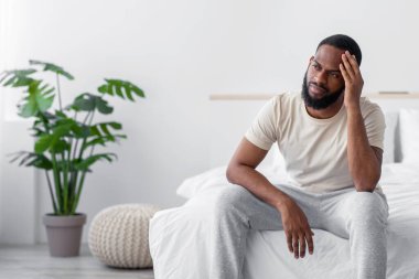 Desperate young african american guy with beard suffers from loneliness, depression in bedroom interior. Problems, stress and self-isolation at home. Difficulty and sad facial expressions, empty space