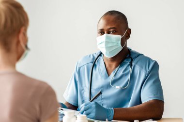Tıbbi personel danışmanlığı, sağlık koruma ve kovid 19 'a karşı aşılama. Koruyucu maskeli ve üniformalı yetişkin Afro-Amerikan doktor klinikte orta yaşlı bir kadınla konuşuyor.