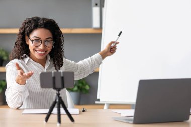 Eğitim, uzaklık çevrimiçi sınıf, görüntülü konuşma ve evde öğrenme. Neşeli bin yıllık Afro-Amerikan kadın öğretmen tahtaya işaret ediyor ve oturma odasındaki akıllı telefon web kamerasına bakıyor.