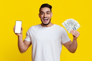 Emotional middle eastern young guy in casual showing smartphone with empty screen and holding bunch of cash over yellow studio background, tradign online on stocks and markets, mockup