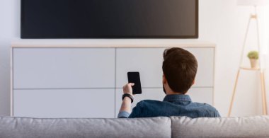 Modern aletler. Akıllı telefonu TV 'ye bağlı, evde video izleyen, arka görüşlü tanınmayan bir adam. Adam telefondan televizyona video aktarıyor. Kablosuz ağ üzerinden aygıtlar arasındaki bilgi aktarımı.