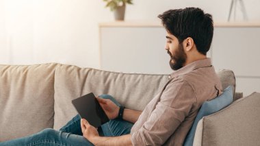 Genç Arap adam dijital tabletle dinleniyor oturma odasında kanepeye yaslanıyor, fotokopi çekiyor. Rahat adam evde eğleniyor, sosyal medyayı kontrol ediyor ya da arkadaşlarıyla sohbet ediyor.