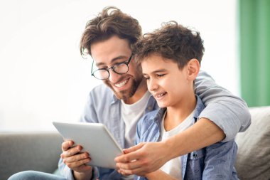 Hafta sonu aile eğlencesi. Mutlu baba ve oğul tablet bilgisayar kullanıyor, internette internette internette geziniyor, rahat koltukta birlikte oturuyorlar. Gadget ve modern yaşam tarzı konsepti