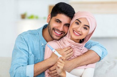 Sevgi dolu Arap çift, pozitif hamilelik testi gösteriyor. Kanepede oturuyor ve kameraya gülümsüyor. Bebek bekleyen genç eşler. Ebeveyn konsepti.