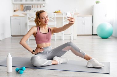 Fitness model blogcusu sosyal ağlar için fotoğraf, evde çalışmak için modern cihaz ve egzersiz yapıyor. Spor giyimli mutlu sarışın bayan mindere oturur oturma odasındaki akıllı telefondan selfie çeker.