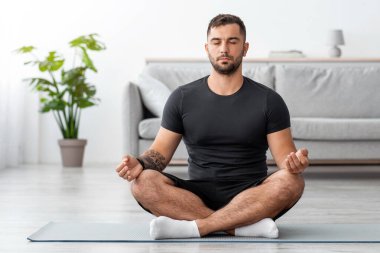 Ciddi şekilde sakin, konsantre olmuş, kaslı, gözleri kapalı beyaz bir adam lotus pozisyonunda oturuyor, oturma odasında yoga yapıyor. Vücut bakımı, meditasyon, karantina süresi dolan barış