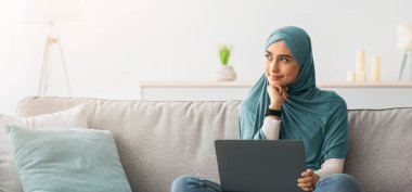 Kompozisyonlar yaz, internetten çalış, sosyal ağlarda sohbet et. Diz üstü bilgisayarı olan neşeli, düşünceli, genç çekici Arap kadın boş alana bakıyor, oturma odasındaki kanepede oturuyor.