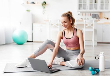 Neşeli beyaz, bin yıllık sarışın fitness kadın blogcusu dizüstü bilgisayara bakıyor, koç sohbeti yapıyor ve evde antreman dersi seçiyor, boş alan. Sosyal uzaklık, aygıtlı spor günlüğü