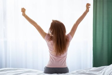 Evde uyandıktan sonra yatağa uzanan genç bir kadının arka görüntüsü. Milenyum bayanı hafta sonu tembelliği yapıyor, kollarını kaldırıyor, uykulu hissediyor, pazar günü evde dinleniyor.