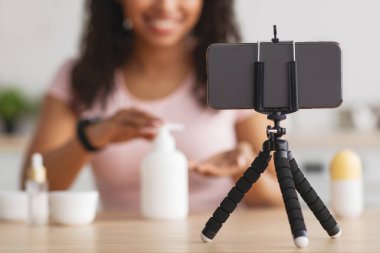 Modern cihazlar, cilt bakımı, kozmetik hakkında güzellik blogu. Mutlu Afrikalı genç Amerikalı kadın krem sıkar ve akıllı telefon webcam 'ine bakar. Üç ayak üzerinde iç kısımdaki masada, seçici bir odak noktası vardır.