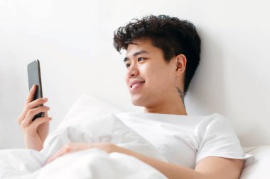 Mobil uygulama, cihazla boş zaman, online reklam ve teklif. Sabah haberleri ve tembellik. Gülümseyen Asyalı genç ya da zoomer, dövmeli beyaz tişörtlü adam akıllı telefona bakar ve yatakta uzanıp, fotokopi çeker.