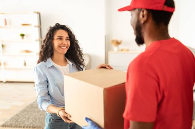 Eve Teslimat Servisi Konsepti. Kapıda bekleyen koruyucu eldivenli Afrikalı Amerikan kurye adamdan büyük bir karton kutu alan gülümseyen genç kadın portresi sipariş ve alışverişleri teslim ediyor.