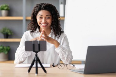 Uzaktan eğitim için modern öğretmen, sınıf dersi, covid-19 salgını ve yeni normaller. Neşeli bin yıllık Afro-Amerikan kadın, arkasında kara tahta, el kol hareketi yapıyor ve akıllı telefon webcam 'ine bakıyor.