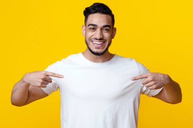 Heyecanlı Arap adam iki elini de boş beyaz tişörtüne gösteriyor ve gülümsüyor sarı stüdyo arka planında poz veriyor. Yakışıklı orta doğulu genç adam elbiselerinde bir şey gösteriyor, boş bir alan.